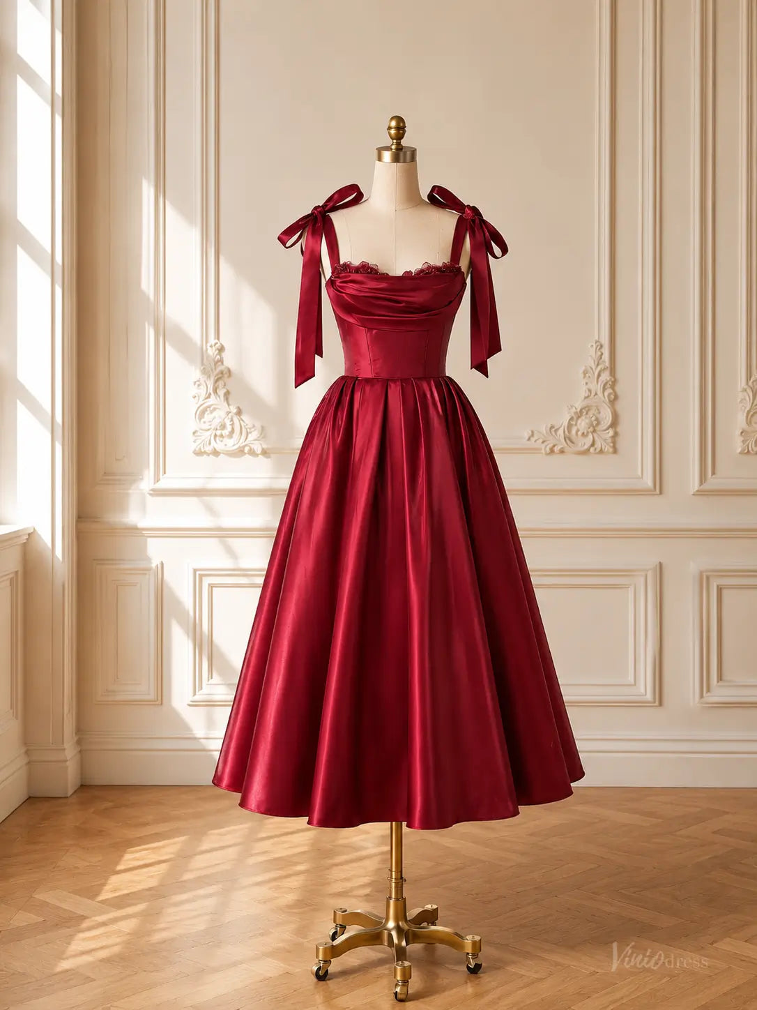 Burgundy satin tea-length prom dress with a corset bodice, draped neckline, and tie bow shoulder straps on a mannequin.