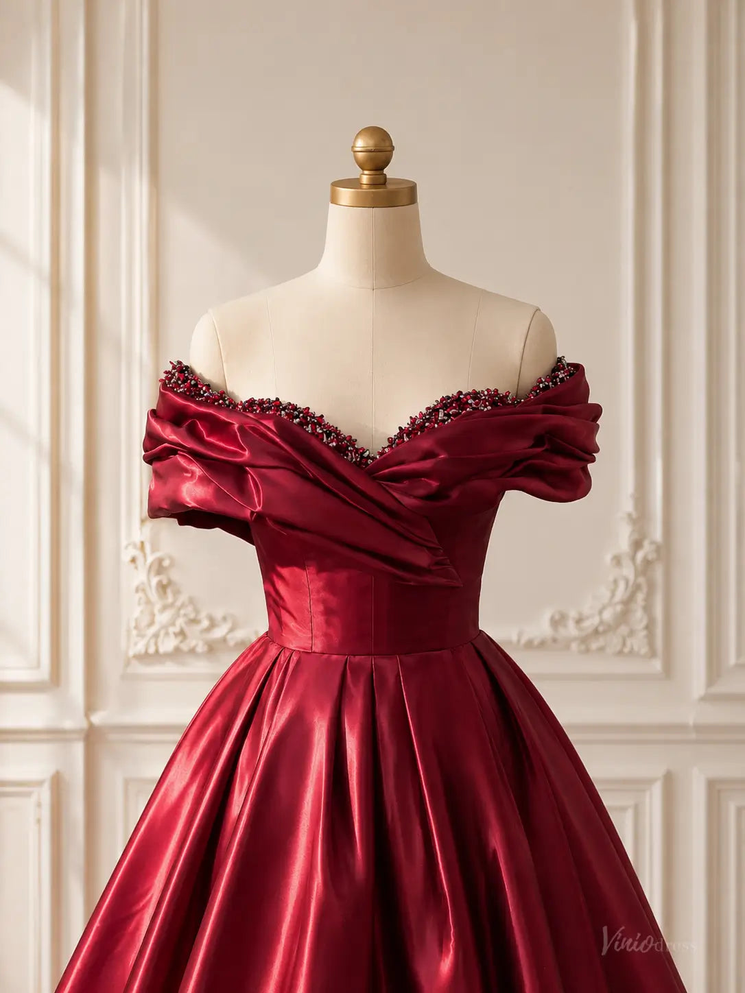 Burgundy satin ball gown with an off-shoulder draped bodice and beaded neckline on a mannequin.