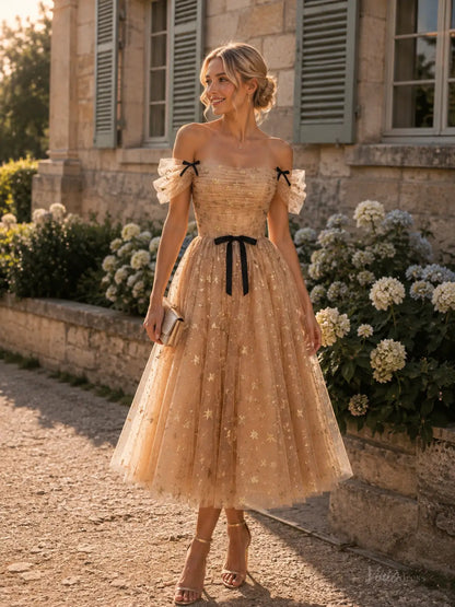 Champagne tea-length tulle prom dress with gold star glitter, off-shoulder sleeves, and black bow accents.