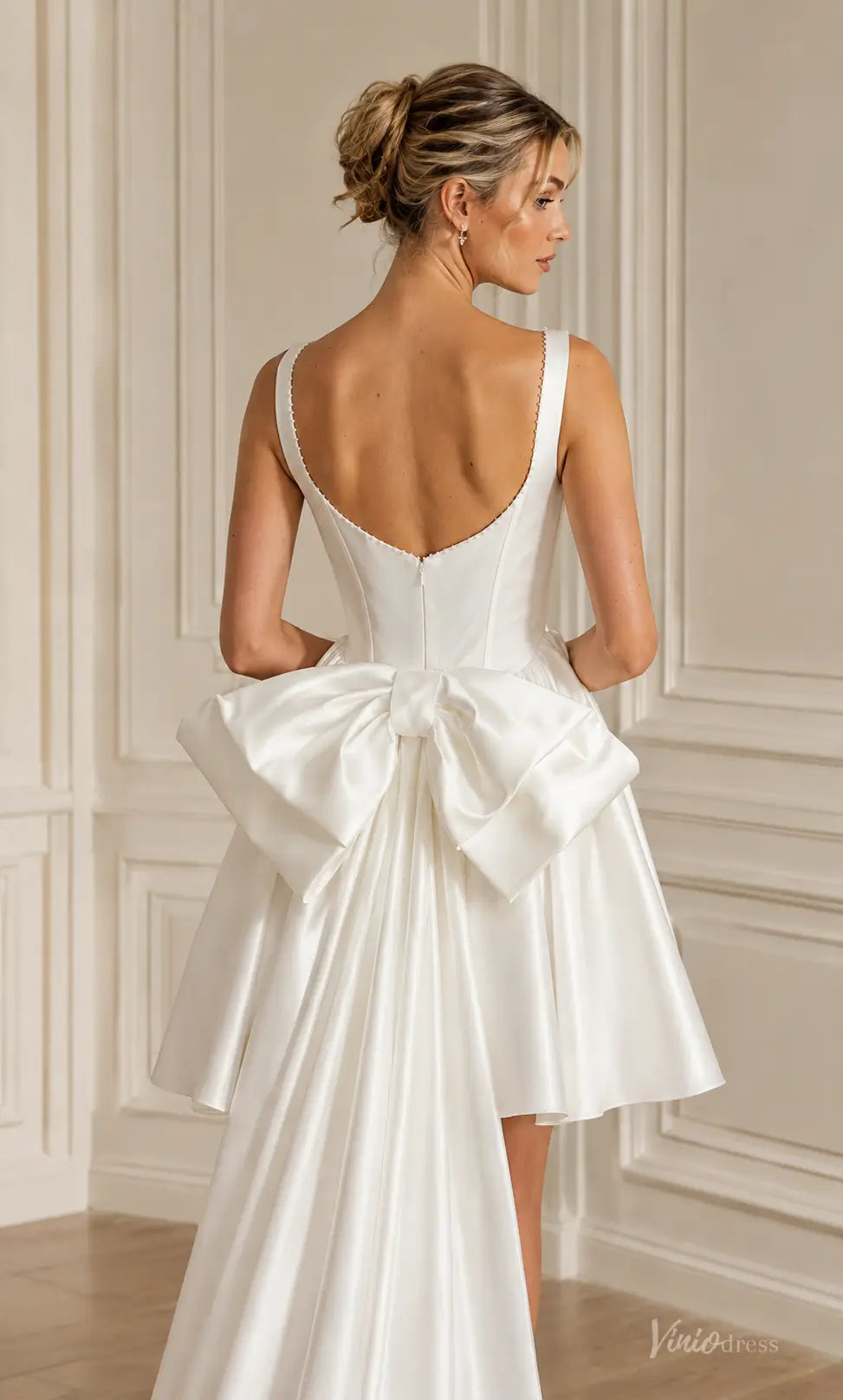 White satin mini wedding dress with a scoop back, sleeveless bodice, and a dramatic oversized bow with a long train.
