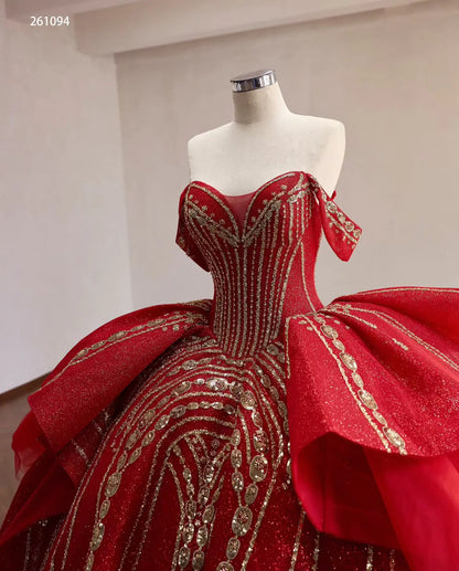Red off-shoulder quinceañera ball gown with gold-beaded corset, sculptural tiered peplum, and glittering fabric.