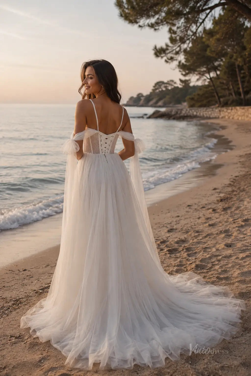 White tulle wedding dress with corset bodice, lace-up back, and detachable off-shoulder sleeves on a beach.