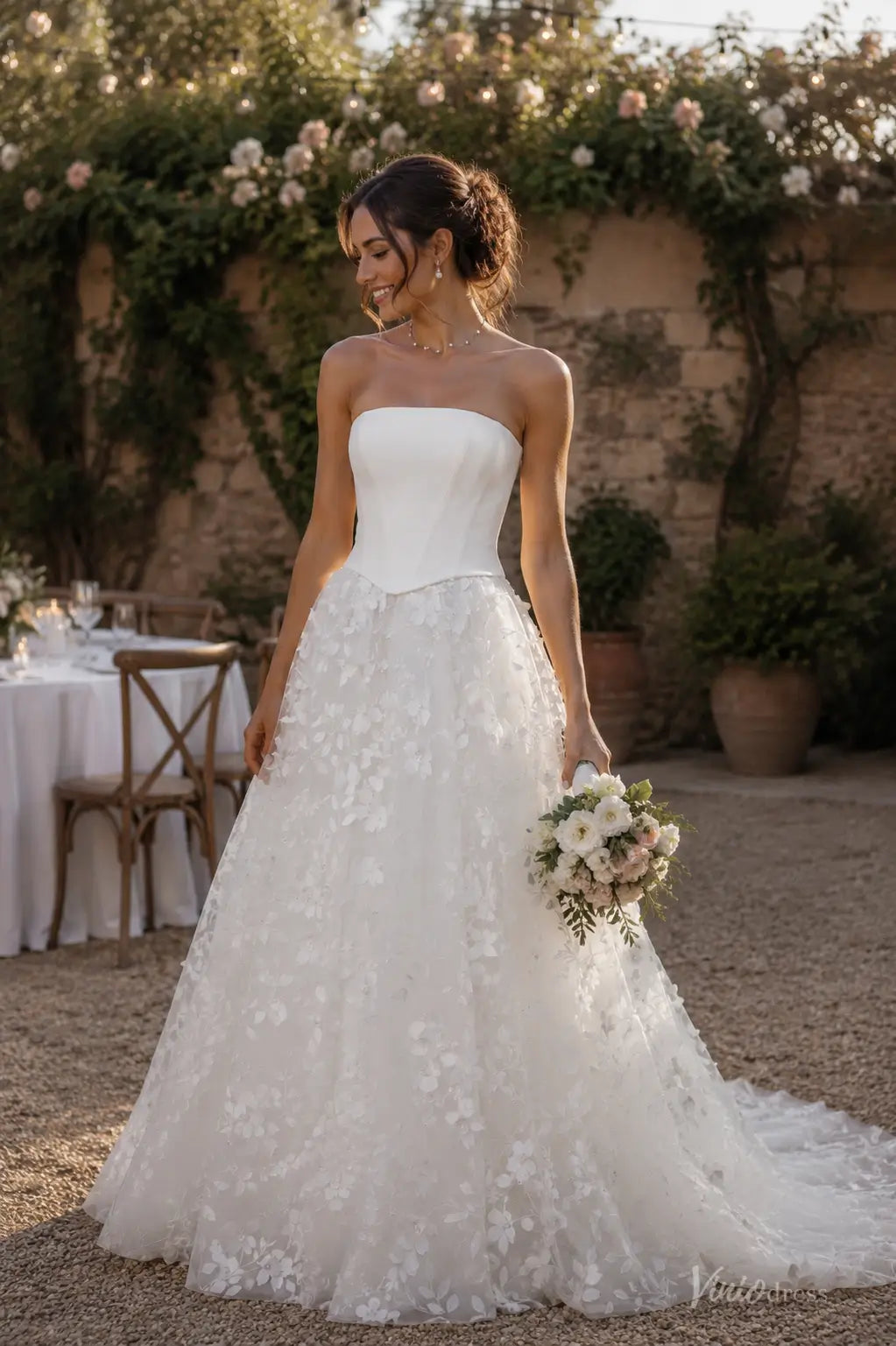 Strapless white wedding dress with a structured bodice and a floral applique A-line skirt, held by a bride outdoors.