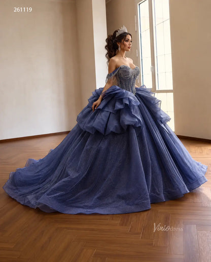 Dusty blue off-shoulder quinceañera dress with a crystal-draped corset, sculptural hip ruffles, and a shimmering full skirt.