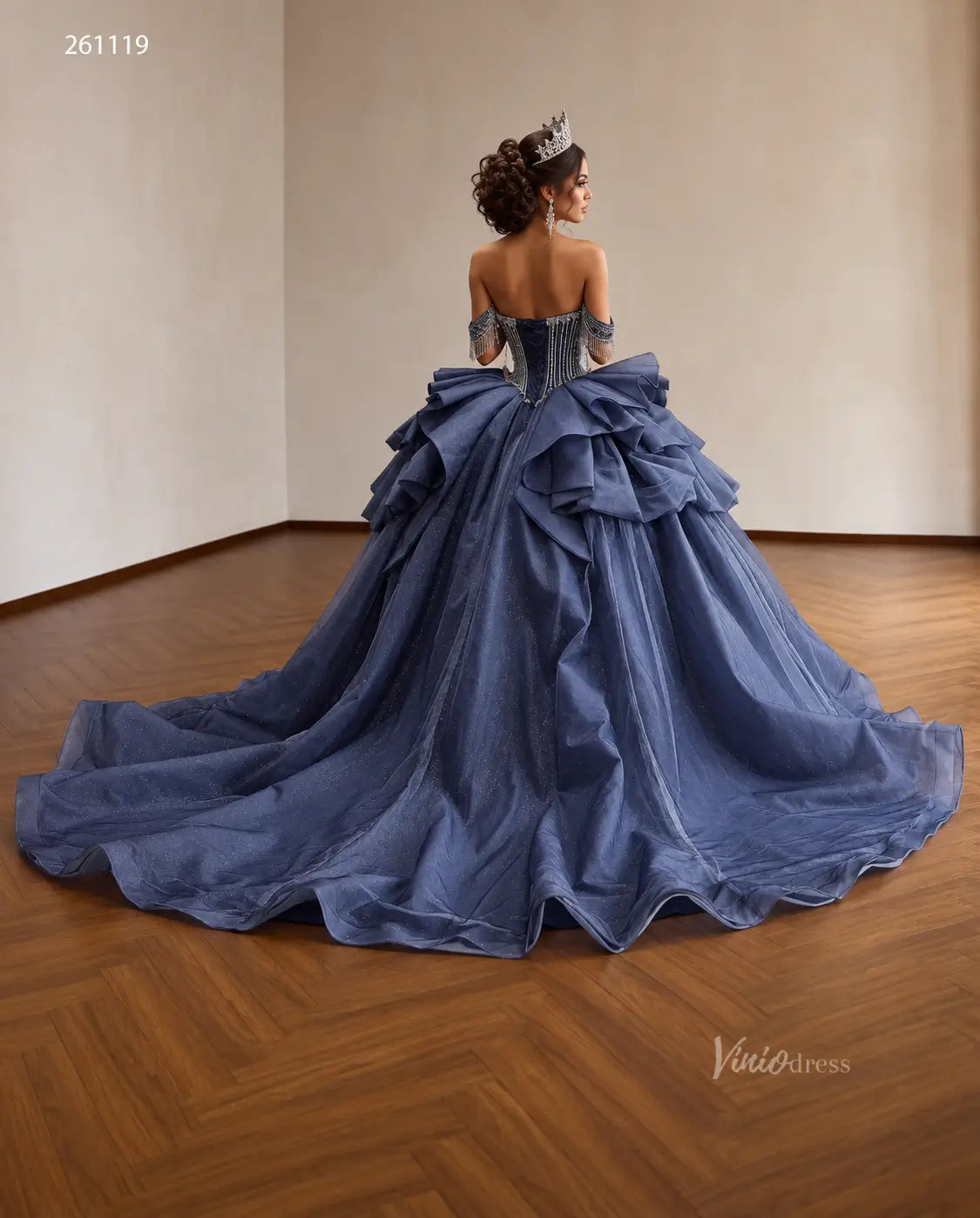 Dusty blue quinceañera dress with crystal-draped corset, off-shoulder straps, and a voluminous ruffled skirt.