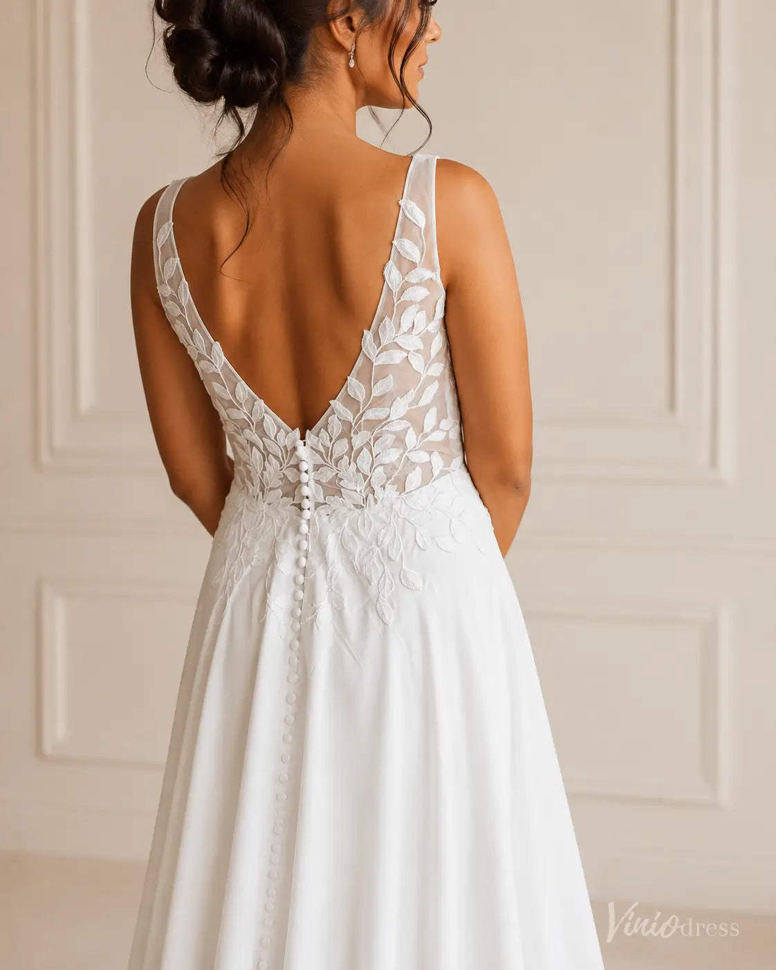 White wedding dress with a deep V-back, sheer lace leaf embroidery, and a delicate row of fabric-covered buttons.
