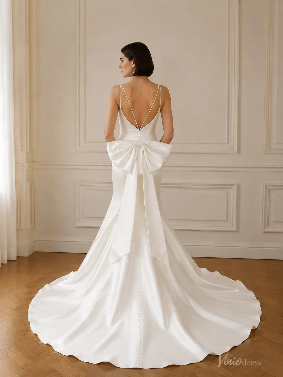 Back view of a white satin mermaid wedding dress with spaghetti straps, a large bow detail, and a button-down train.