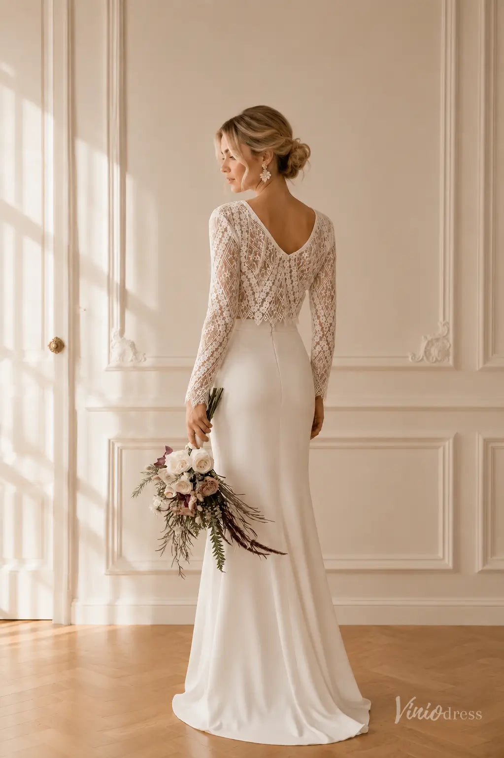 Boho two-piece wedding dress with long-sleeve lace top and sleek white crepe sheath skirt, held by a bride with a bouquet.