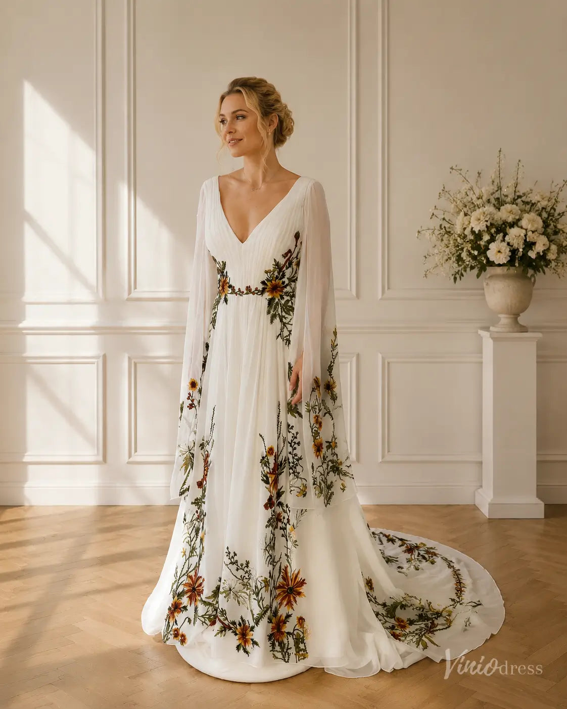 White chiffon wedding dress with V-neck, flowing cape sleeves, and colorful meadow floral appliqués on a long skirt.