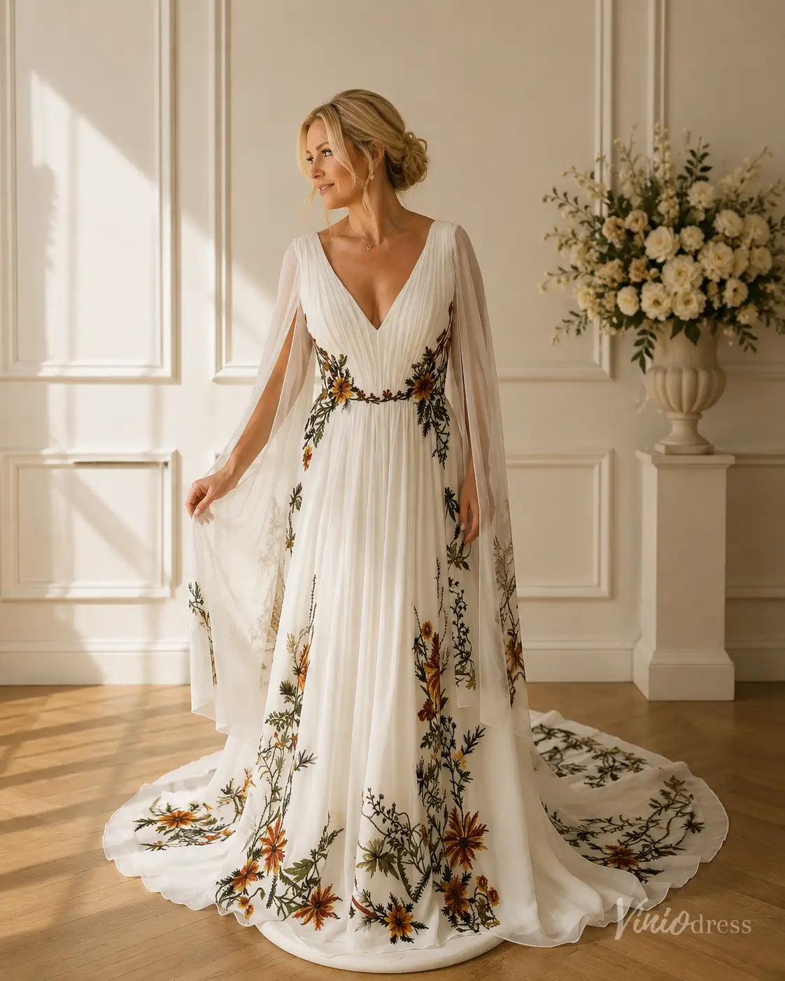 White chiffon wedding dress with V-neck, flowing cape sleeves, and colorful meadow floral appliqués on a full skirt.